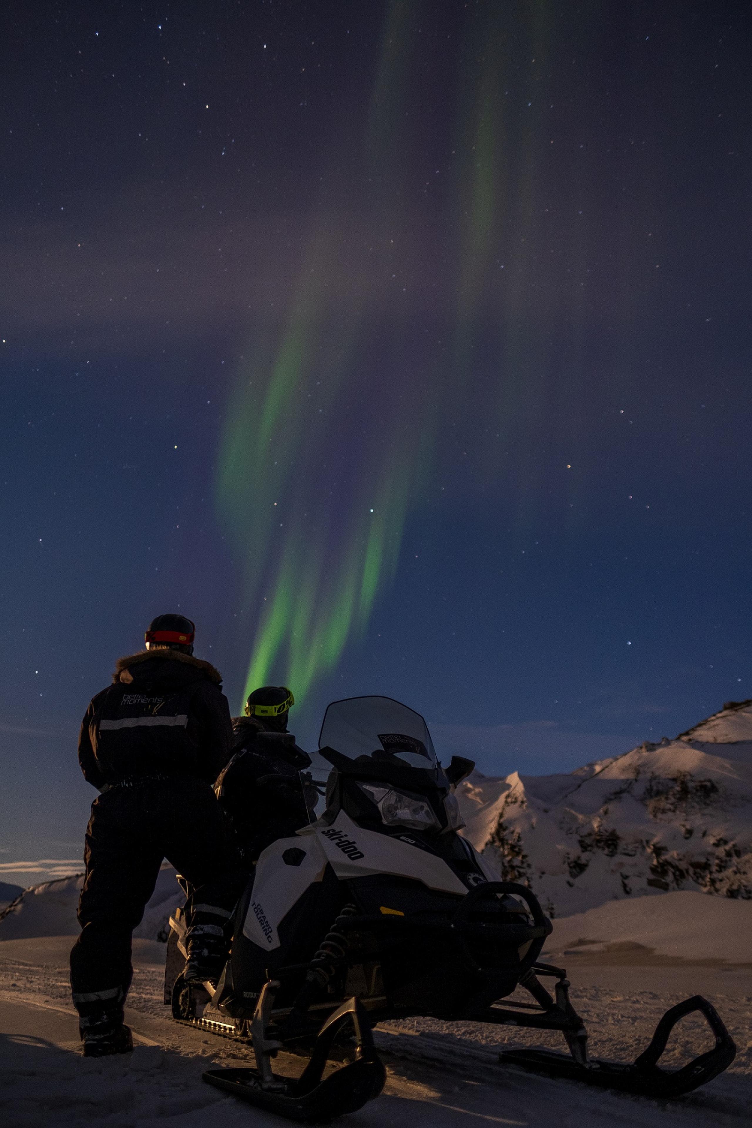 To personer som står bak en snøscooter og ser på nordlys på himmelen
