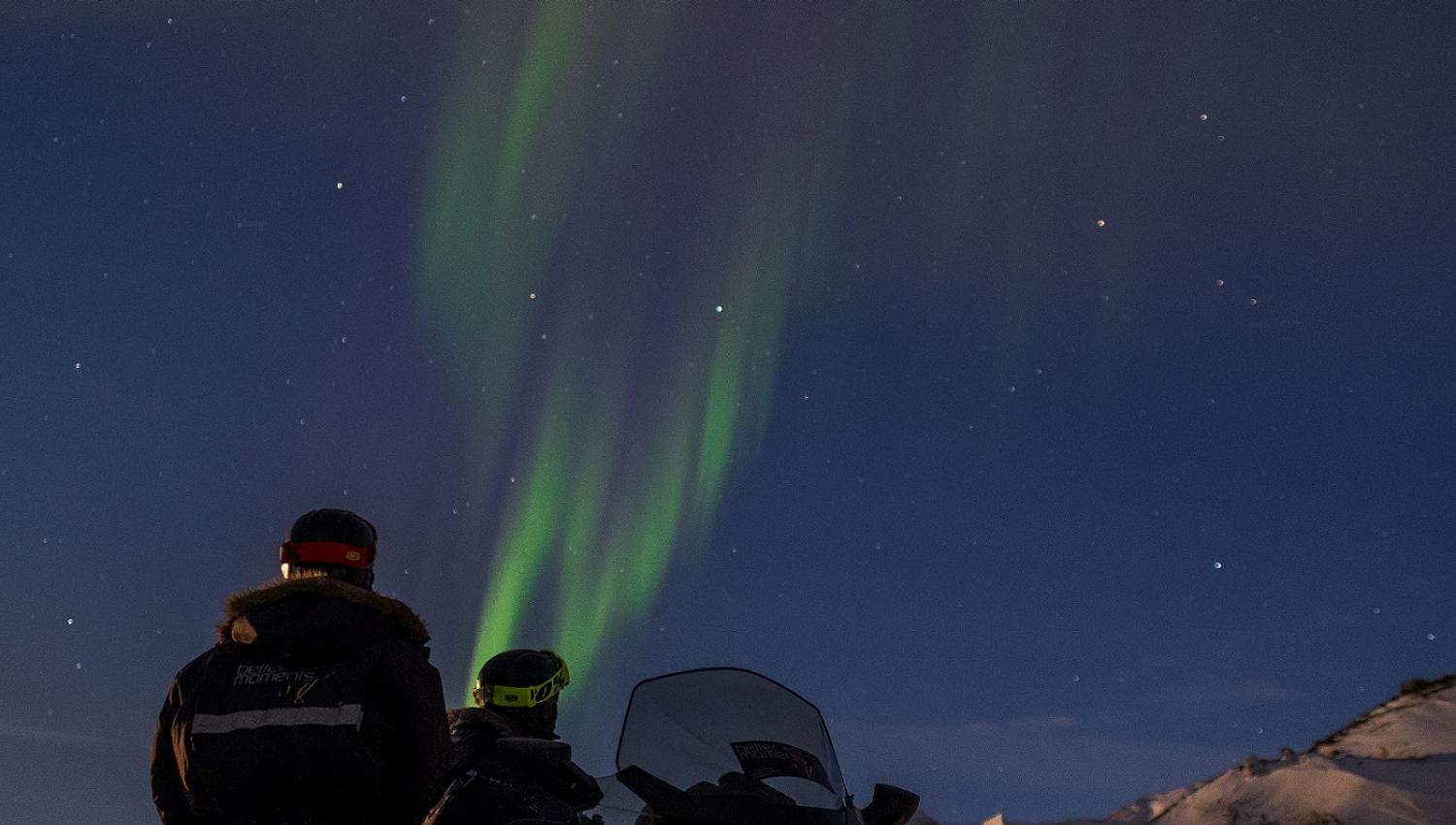 Two persons standing behind a snowmobile and looking up at northern lights in the sky