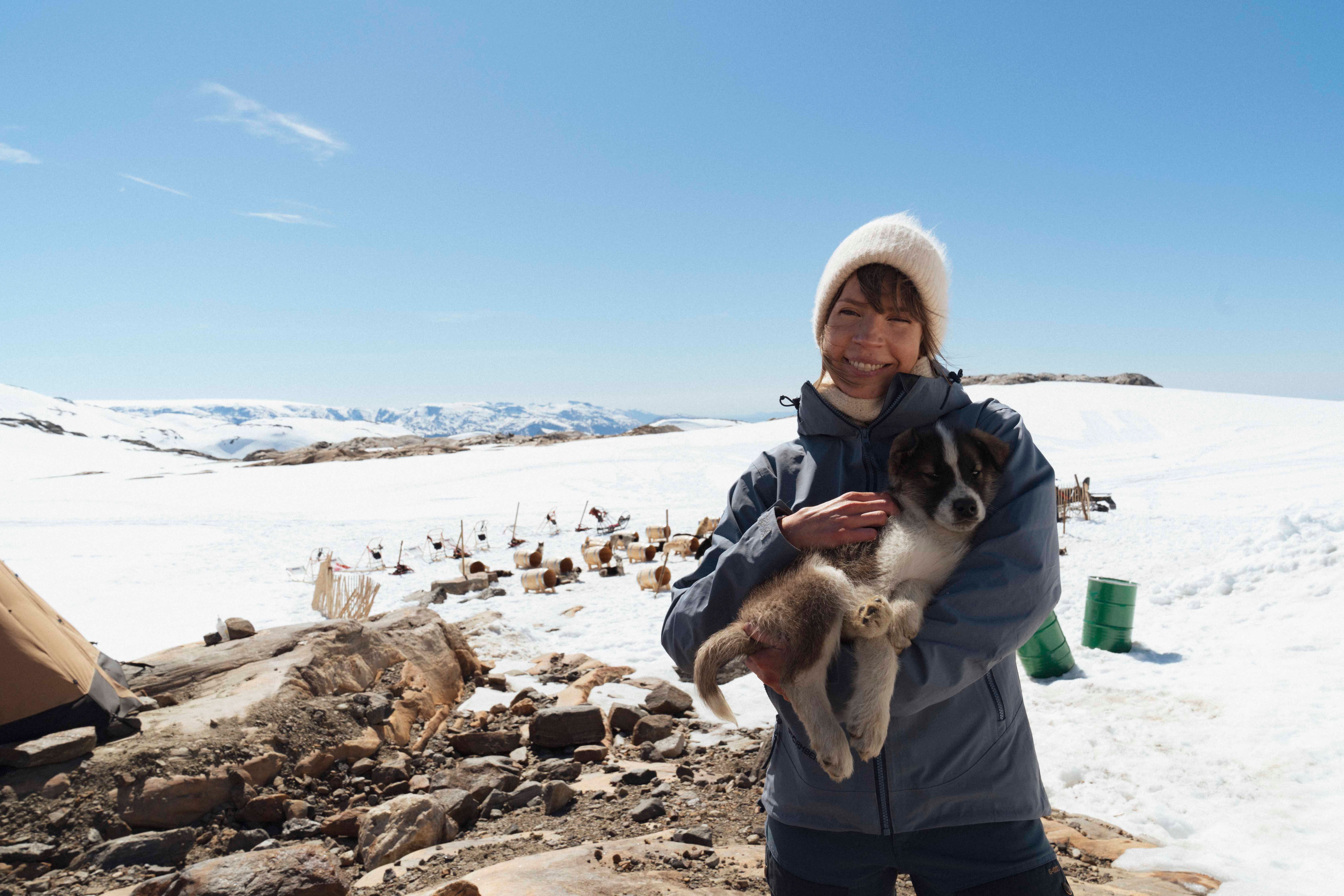A person smiling and holding a huskypuppy with folgefonna husky camp and mountains in the background.