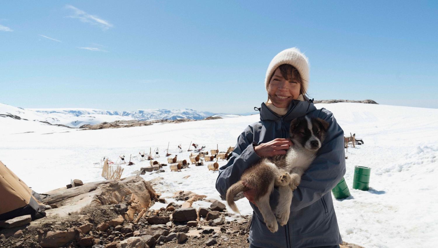 A person smiling and holding a huskypuppy with folgefonna husky camp and mountains in the background.