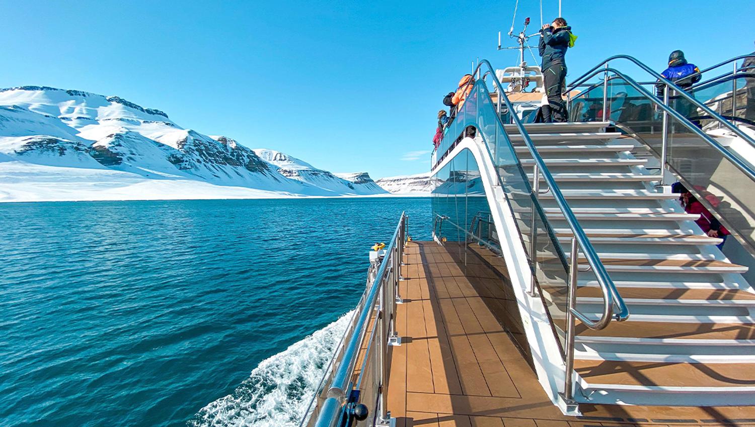 A boat sailing through a fjord with guests on board and snow-covered mountains in the background