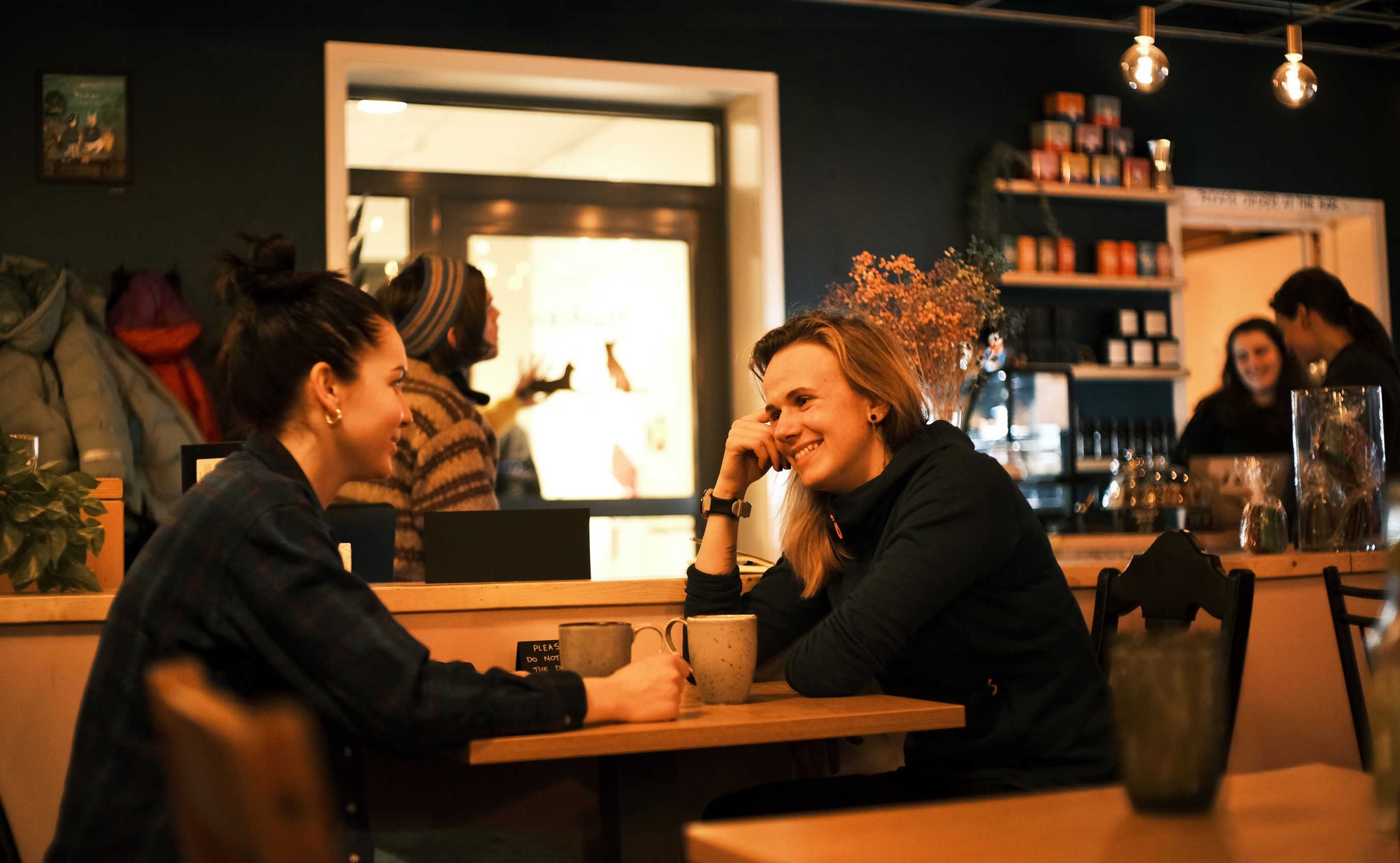 Two guests sitting at a table drinking coffee together, and employees in the background behind the café counter.