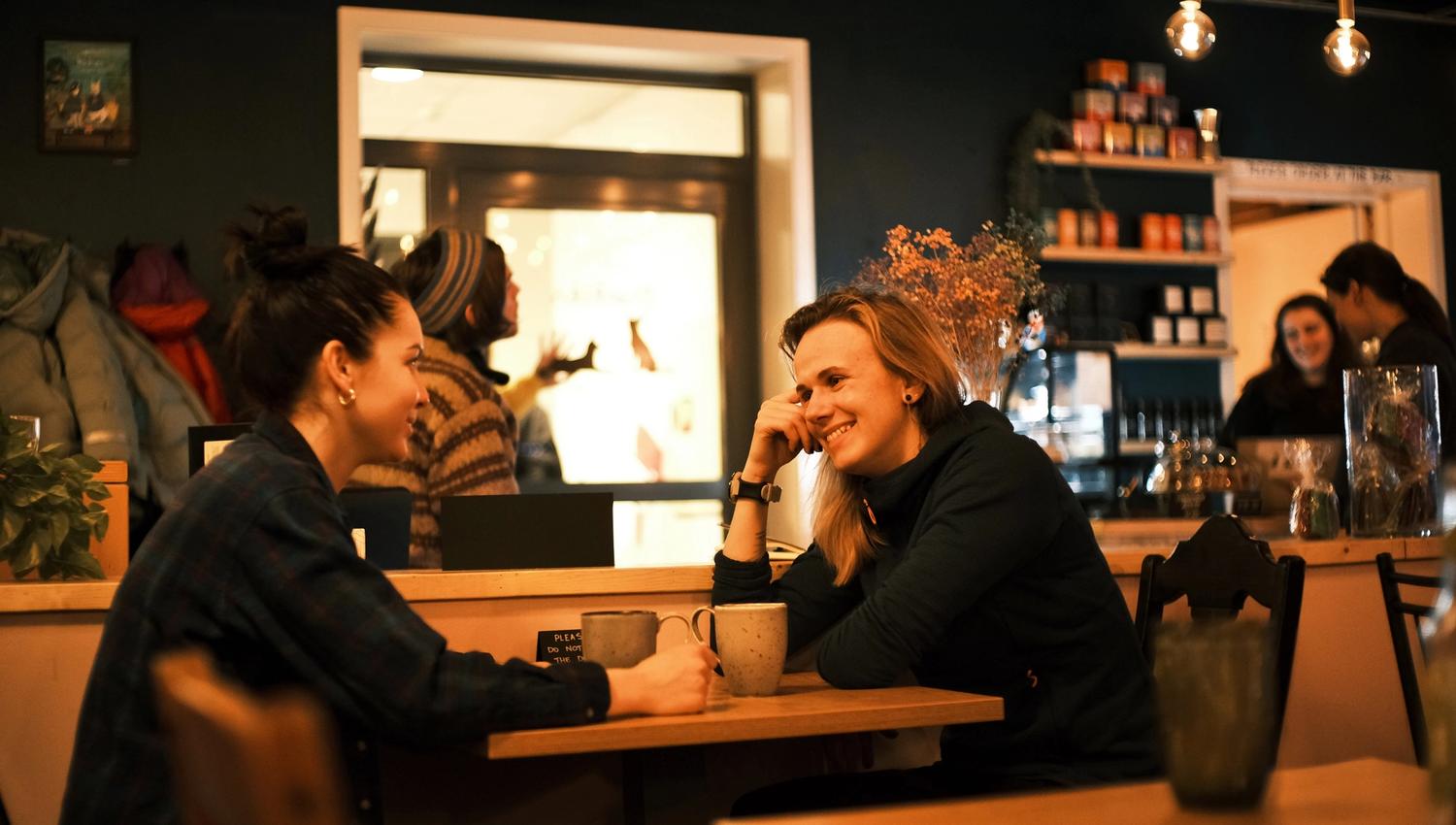 Two guests sitting at a table drinking coffee together, and employees in the background behind the café counter.