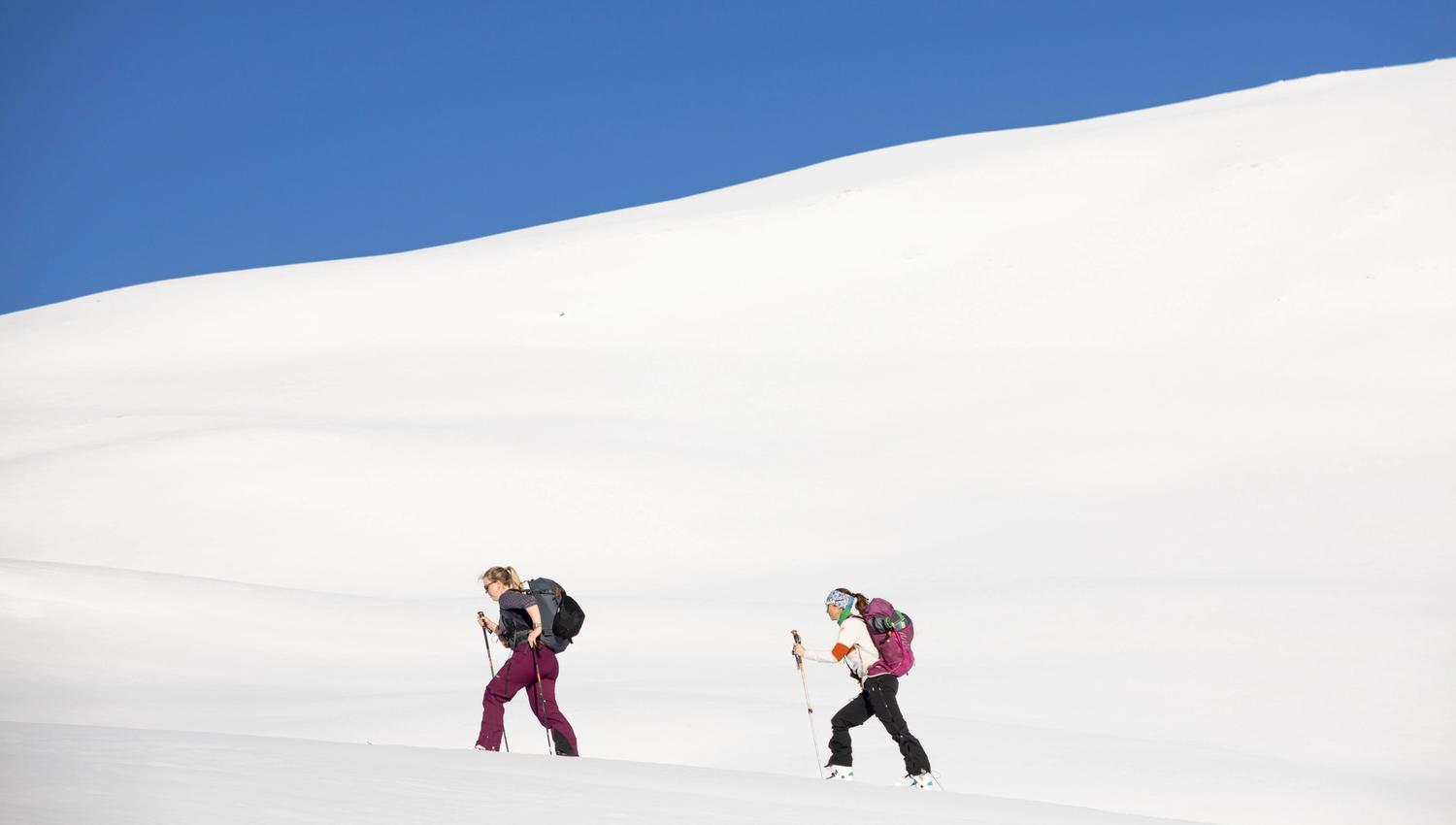 Two female skiers wearing backpacks walking uphills.