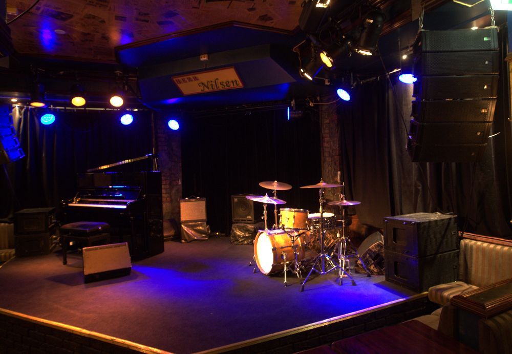 Stage with drum kit, piano, various amplifiers and speakers.