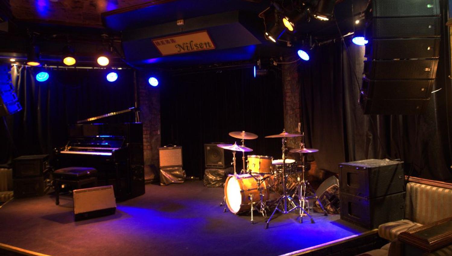 Stage with drum kit, piano, various amplifiers and speakers.