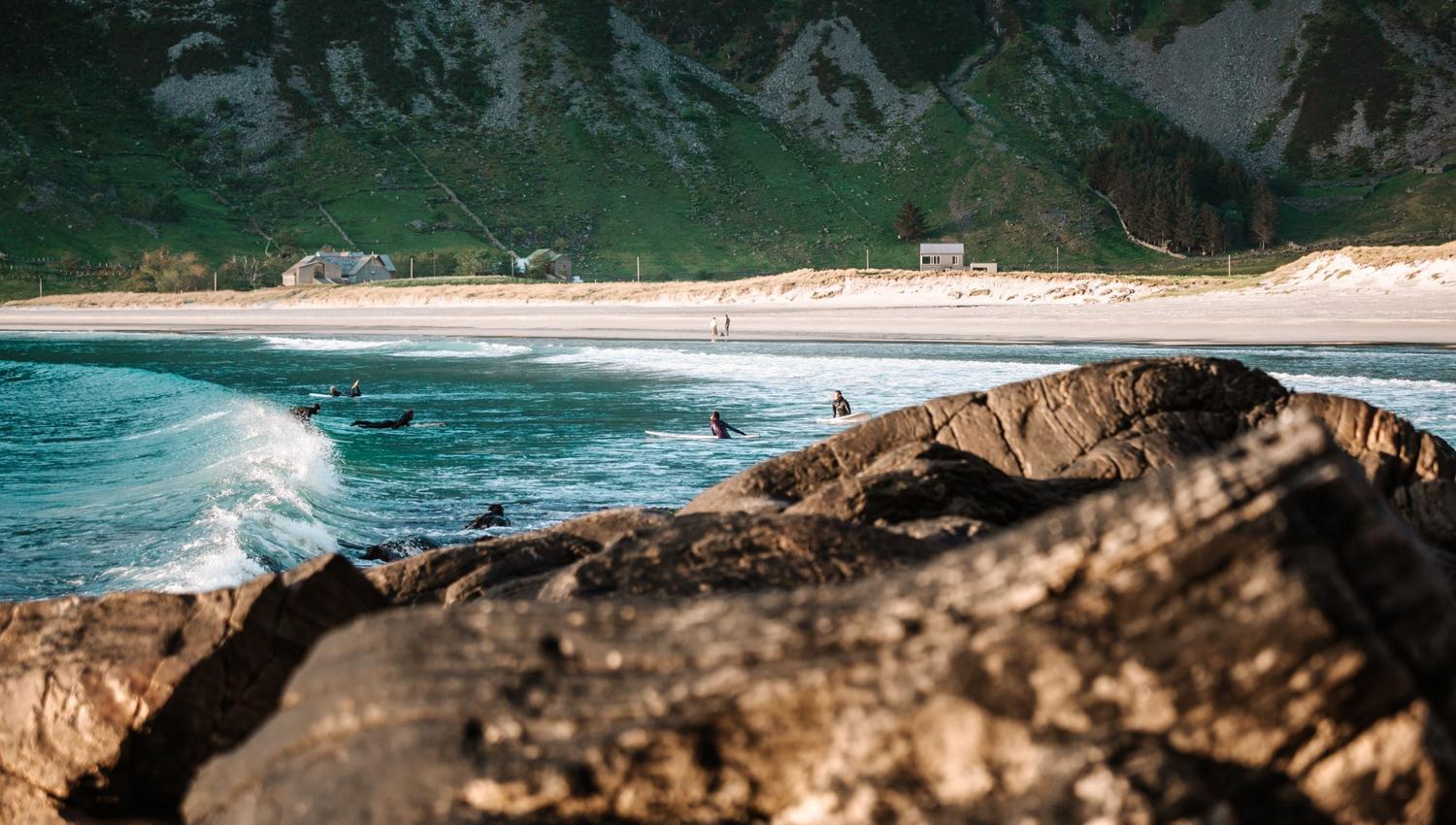 Surfaranar i bølgjene nær store steinar, med sandstrand, små hus og grøne fjell i bakgrunnen..