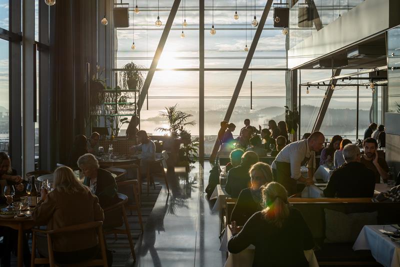 People dining in a modern restaurant with large panoramic windows and a sunset view over the Oslofjord