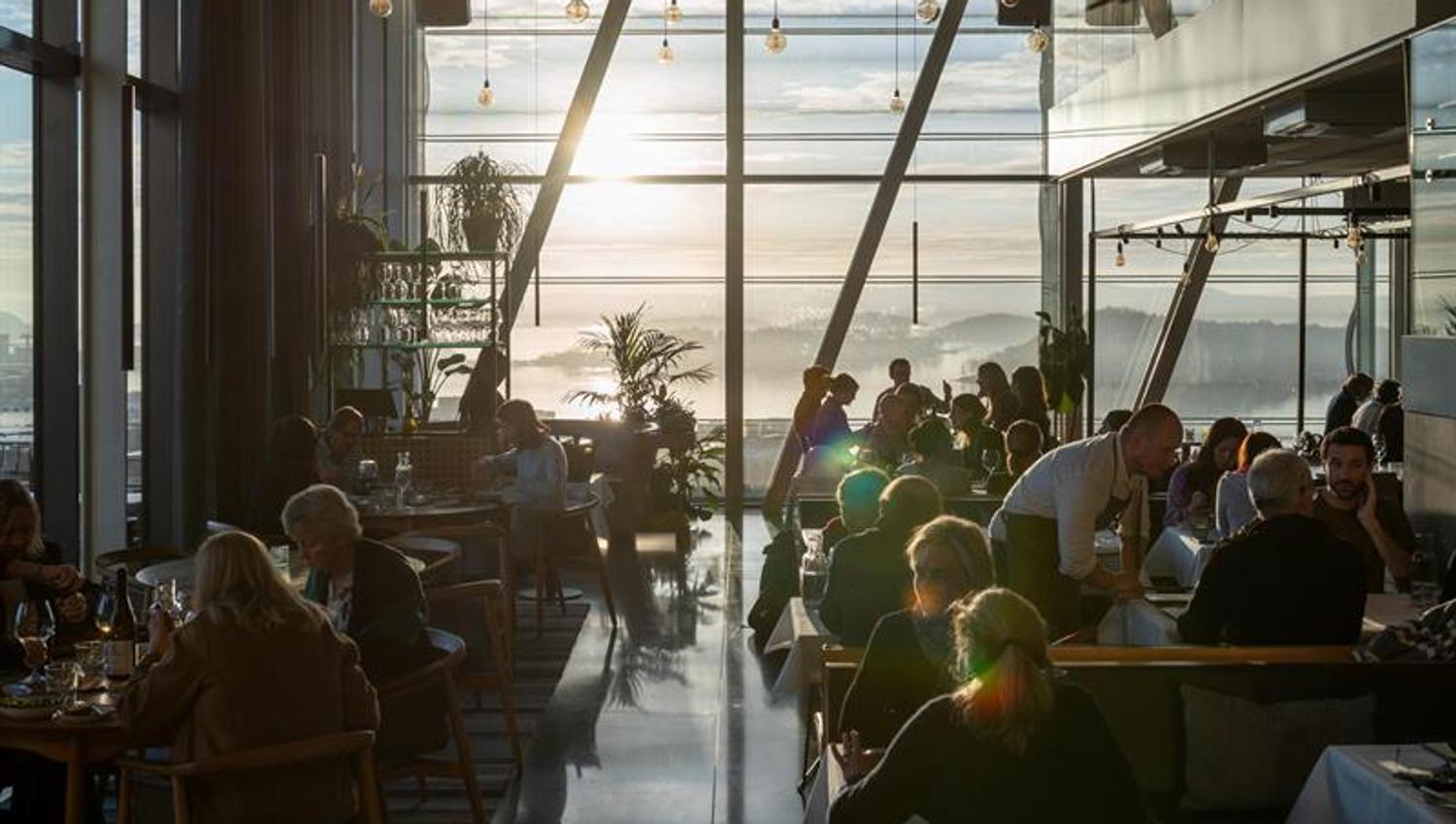 People dining in a modern restaurant with large panoramic windows and a sunset view over the Oslofjord