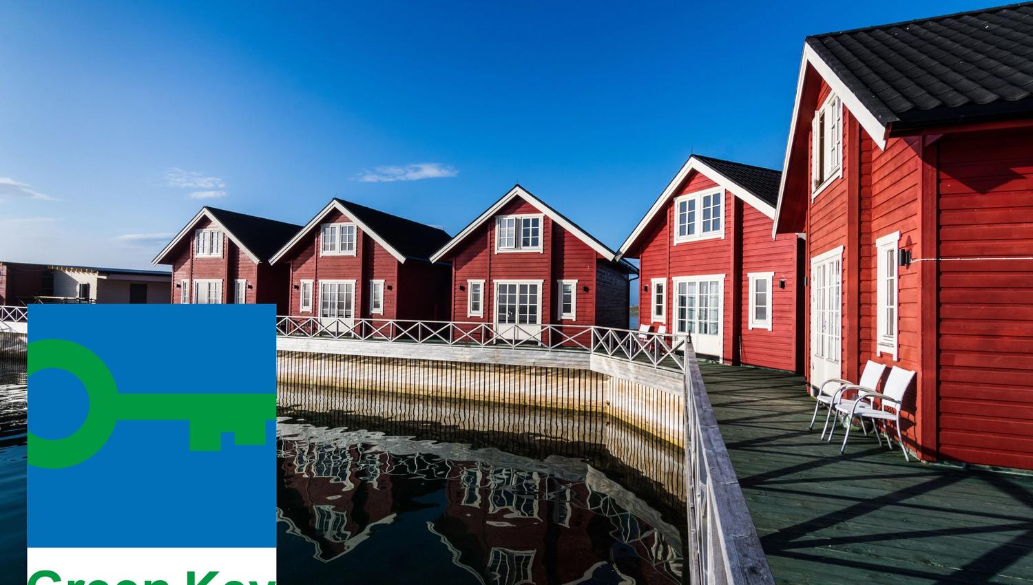 Red wooden houses with white details by the waterfront, reflected in the water. 'Green Key' logo in the bottom left corner