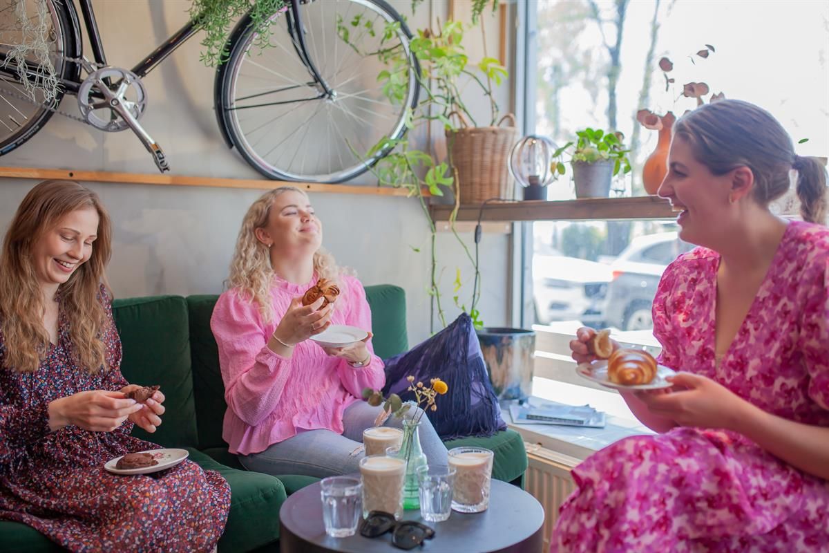 Three people sit at a café window table, enjoying food and drinks in a bright, modern setting.
