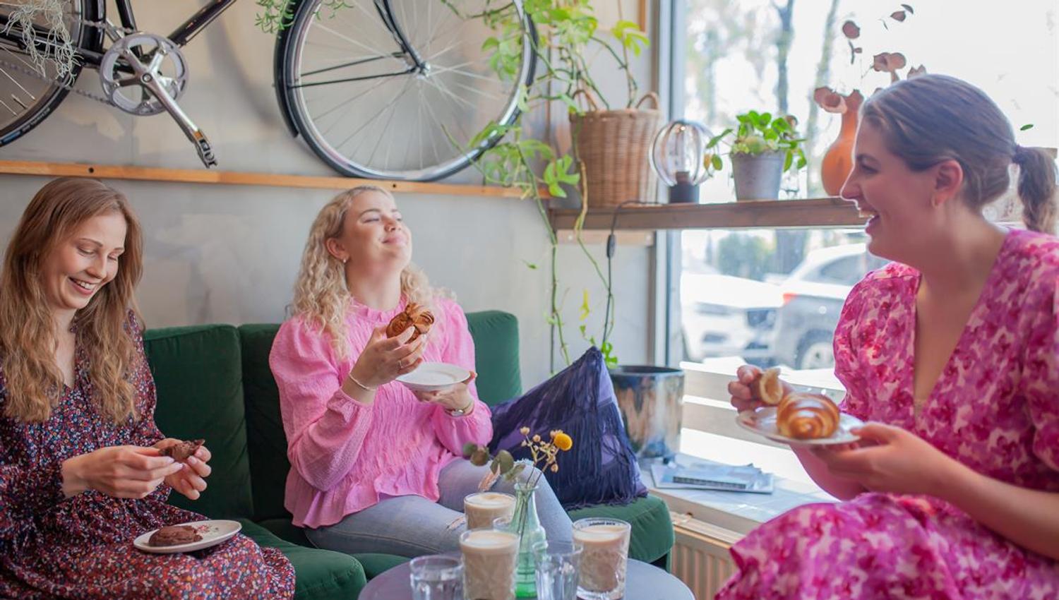 Three people sit at a café window table, enjoying food and drinks in a bright, modern setting.