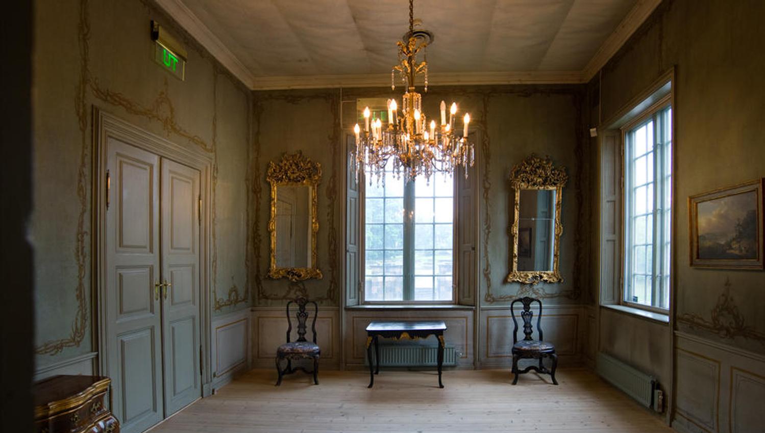 Old interior with a chandelier hanging from the ceiling, mirrors and paintings on the walls.