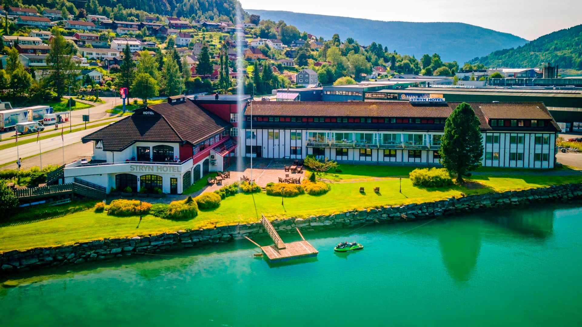 Stort hotellbygg ved grøn plen og kai ved turkist fjordvatn, med skogkledde åsar og bustader i bakgrunnen..