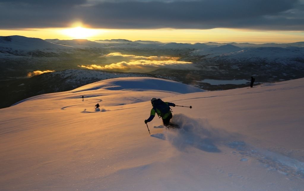 A skier descends a snowy slope from Hestefjellet in Stryn at sunset, kicking up powder, with mountains and low clouds in the distance..