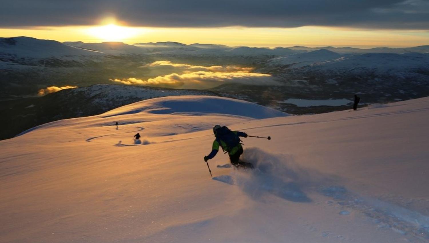 A skier descends a snowy slope from Hestefjellet in Stryn at sunset, kicking up powder, with mountains and low clouds in the distance..