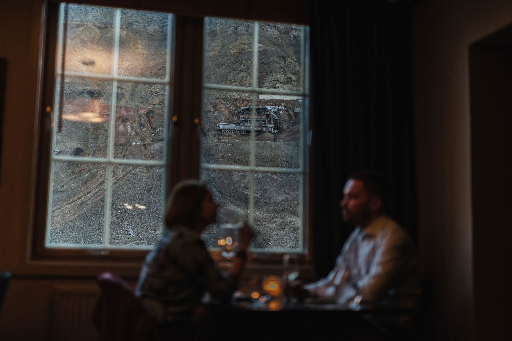 Two persons sitting at a restaurant table with a view towards a mountain and an abandoned mine through a window in the background