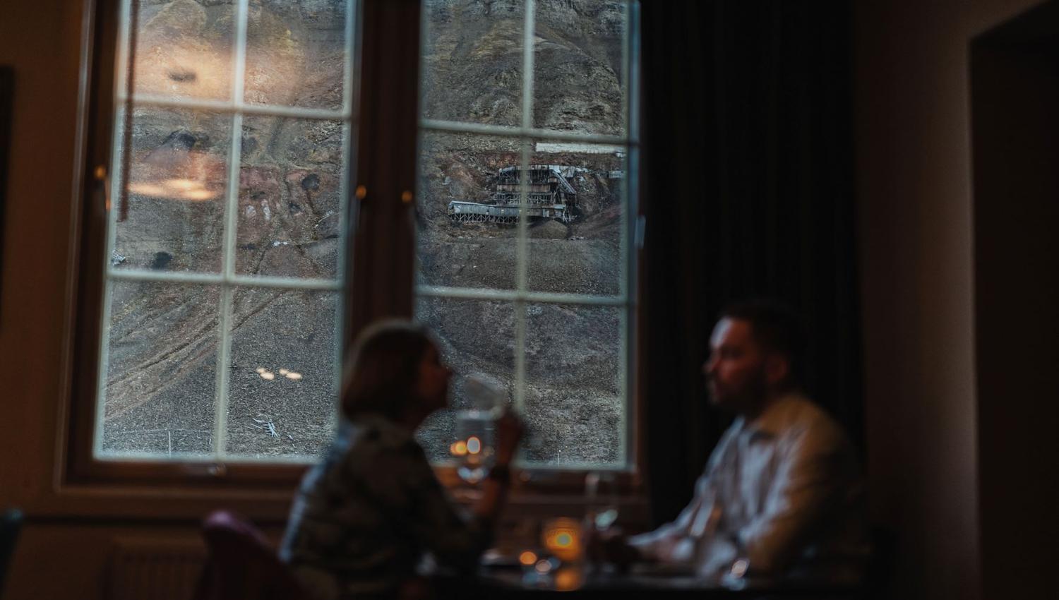 Two persons sitting at a restaurant table with a view towards a mountain and an abandoned mine through a window in the background