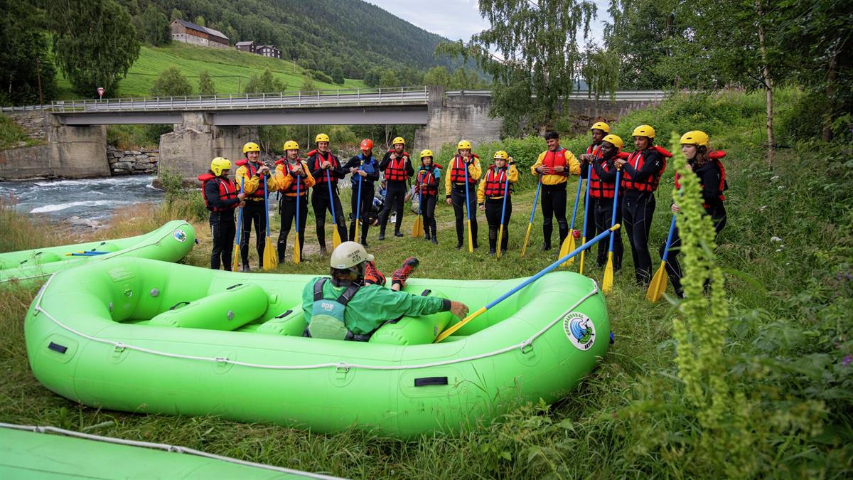 Group with rafting equipment gathered around a green raft at the riverbank, with a bridge and valley in the background.