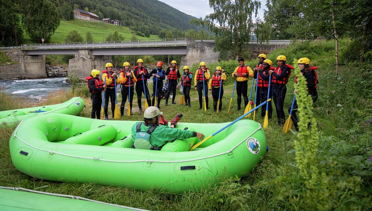 Group with rafting equipment gathered around a green raft at the riverbank, with a bridge and valley in the background.