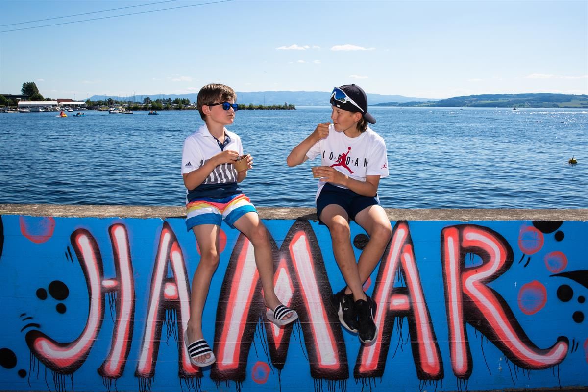 Two people sit by the waterfront in front of a large colorful “Hamar” sign, with Lake Mjøsa in the background.