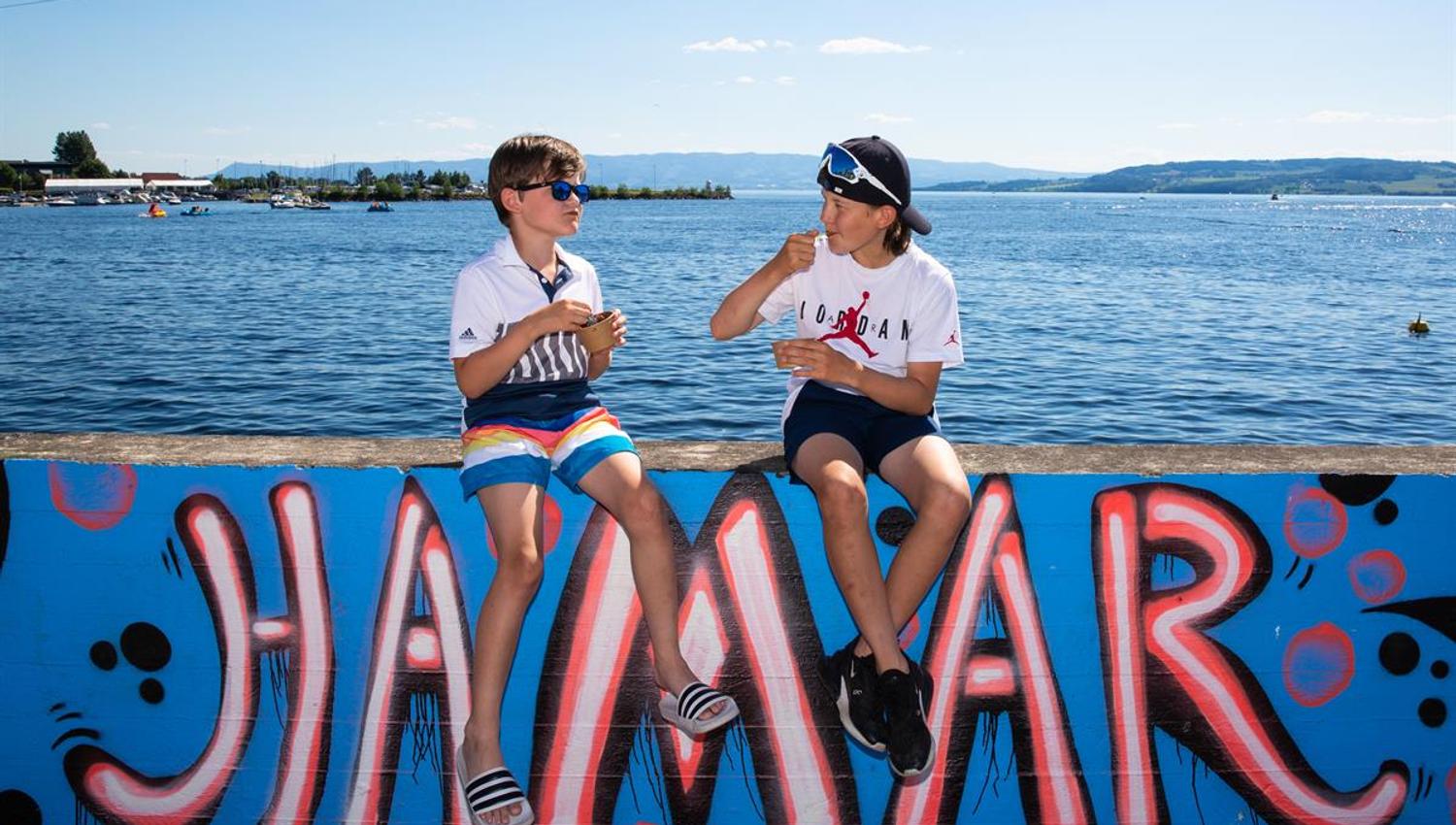 Two people sit by the waterfront in front of a large colorful “Hamar” sign, with Lake Mjøsa in the background.