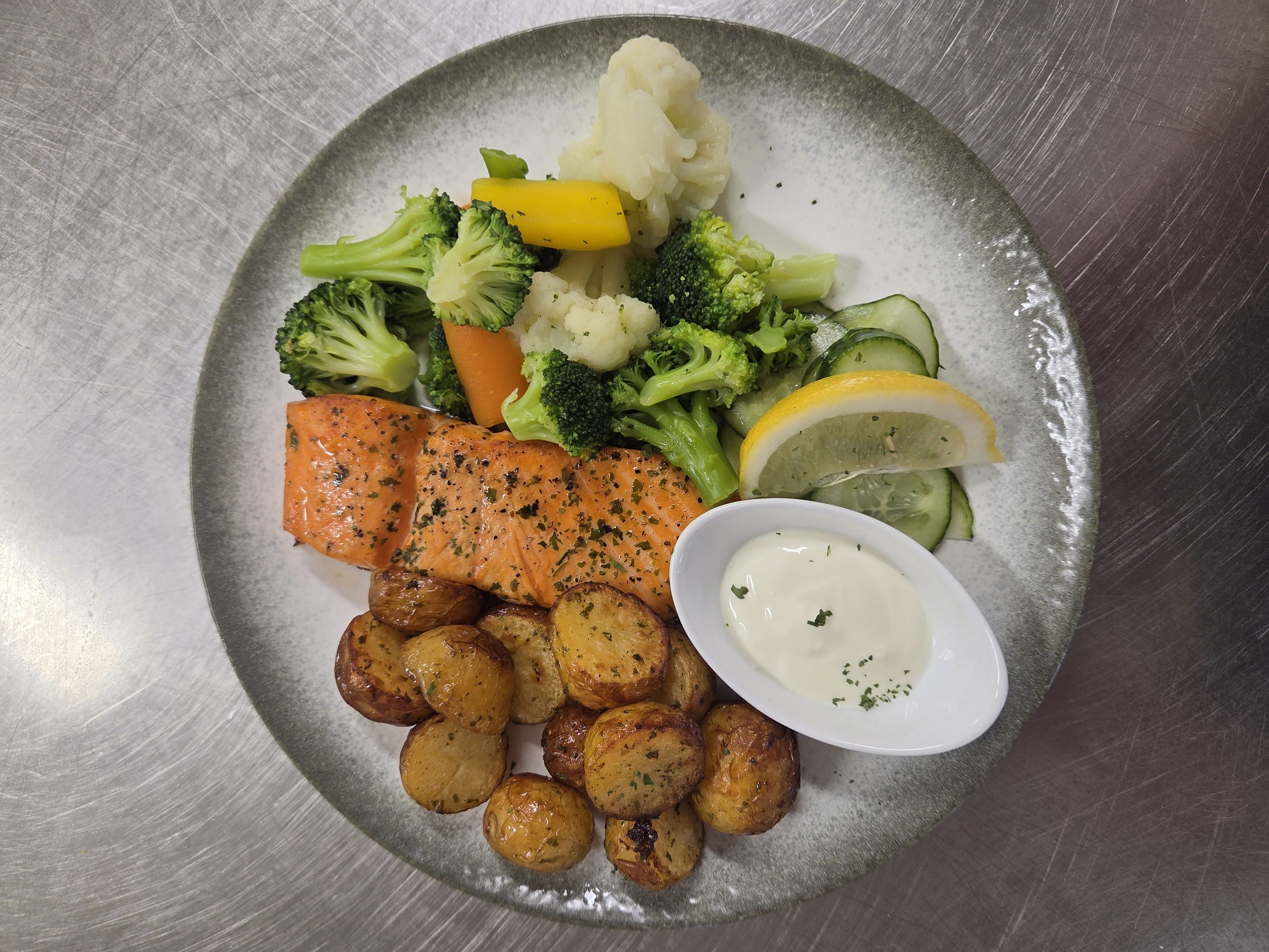 Overhead view of a plate with herb-seasoned salmon, roasted potatoes, mixed vegetables, lemon, cucumber, and sauce.