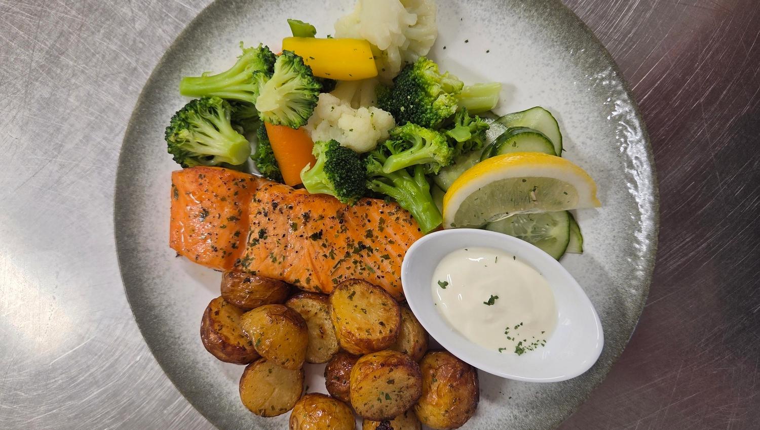 Overhead view of a plate with herb-seasoned salmon, roasted potatoes, mixed vegetables, lemon, cucumber, and sauce.