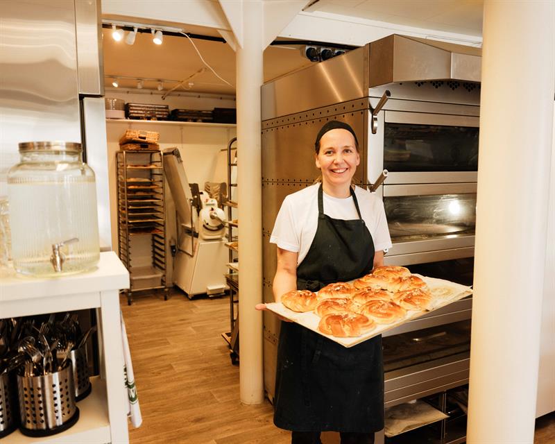 A baker stands inside Iversen Bakery and Gallery holding a tray of freshly baked buns, surrounded by ovens, baking equipment and work surfaces.