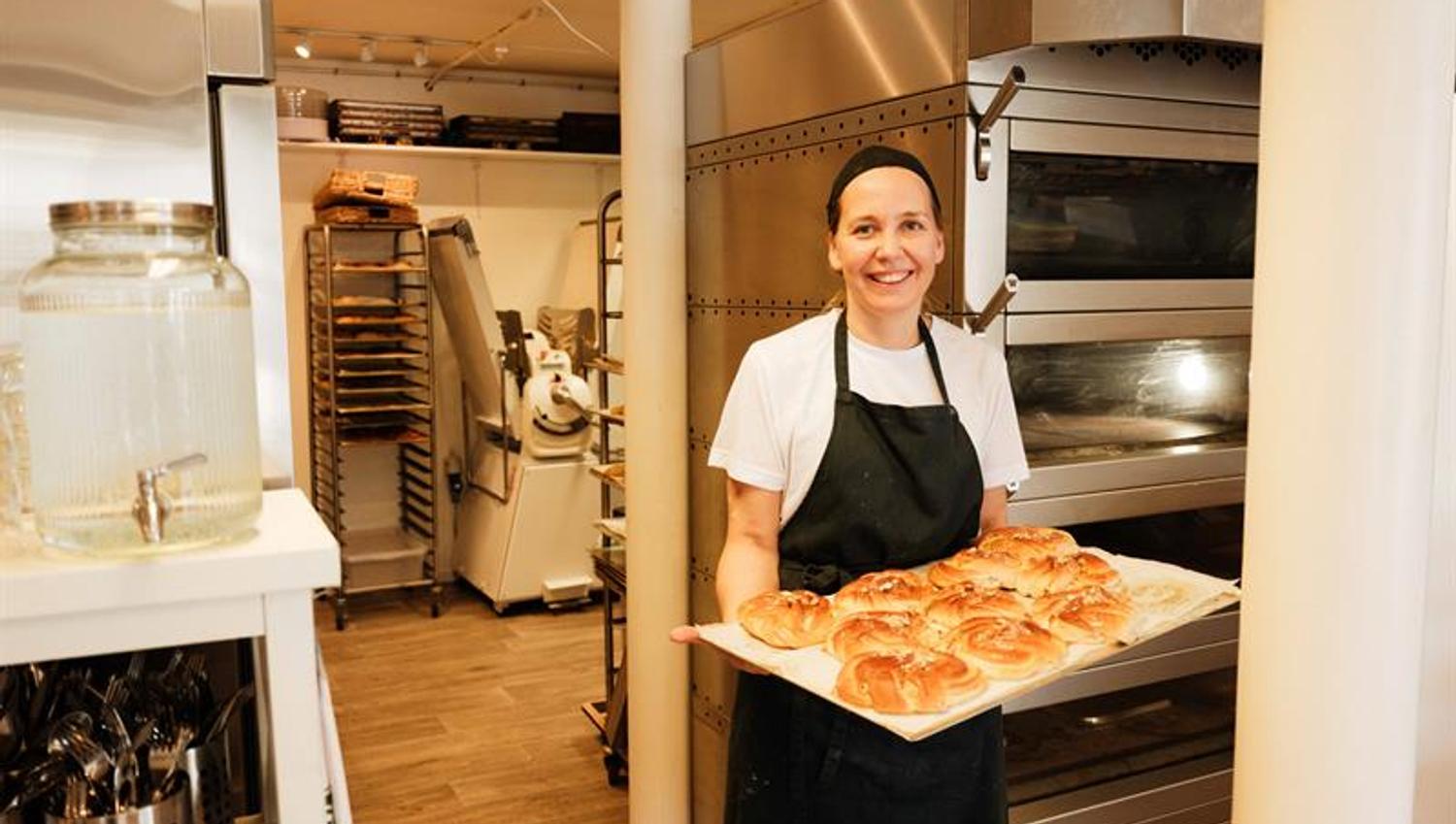 A baker stands inside Iversen Bakery and Gallery holding a tray of freshly baked buns, surrounded by ovens, baking equipment and work surfaces.