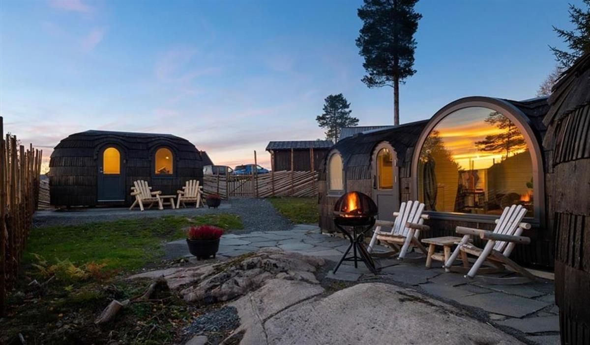 An outdoor area with wooden cabins, seating, and a lit firepit at dusk.