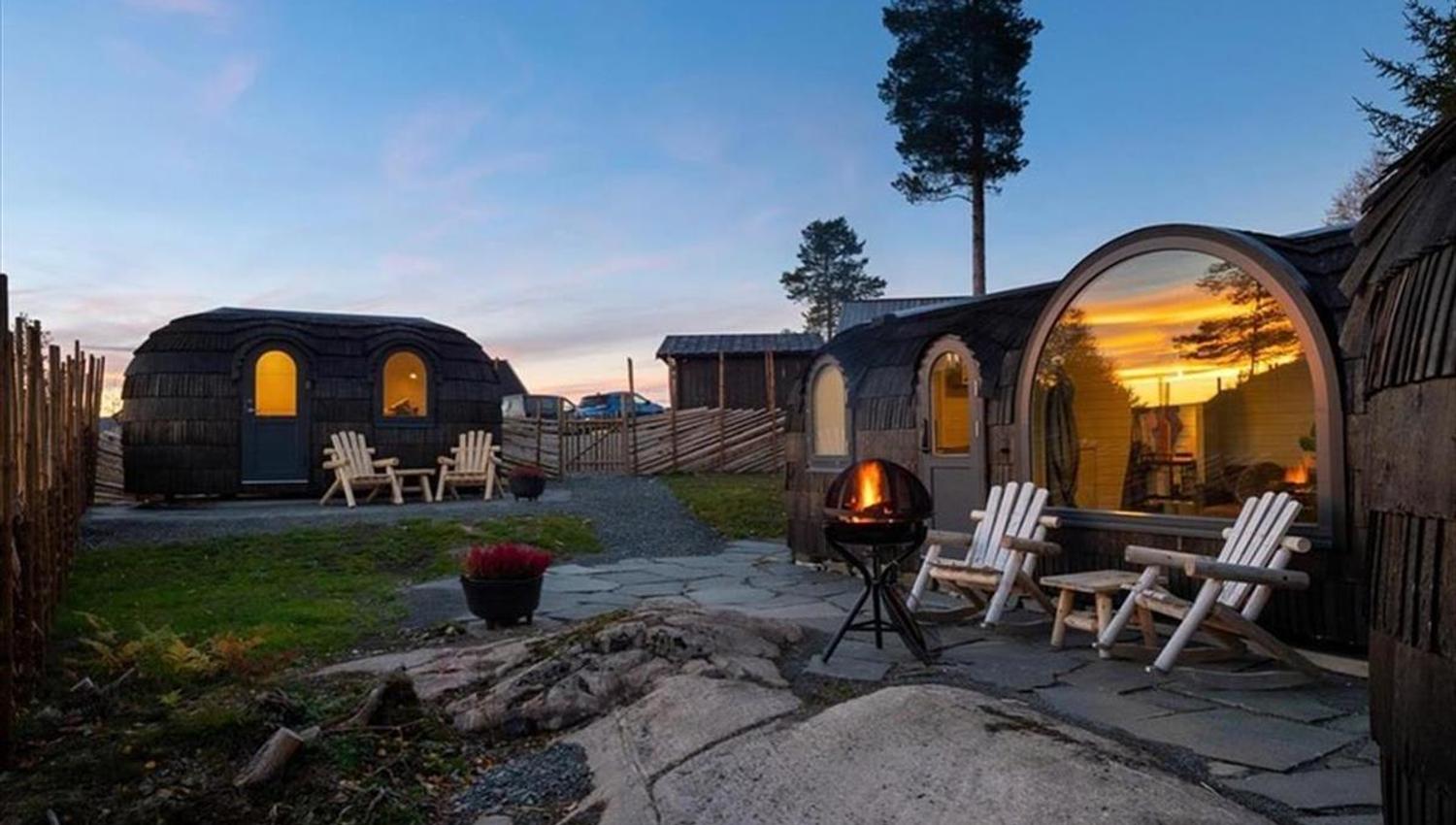 An outdoor area with wooden cabins, seating, and a lit firepit at dusk.