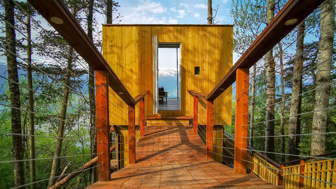 A wooden bridge with cable rails leads to a small timber cabin among trees, with lake and mountains beyond..