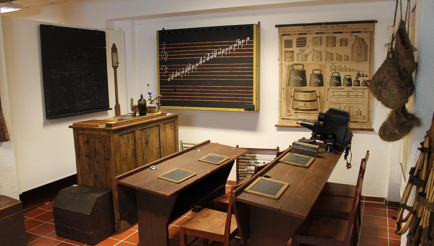 Classroom with many different details. Brown chairs and desks, blackboard, notice board, posters, bags, chest.