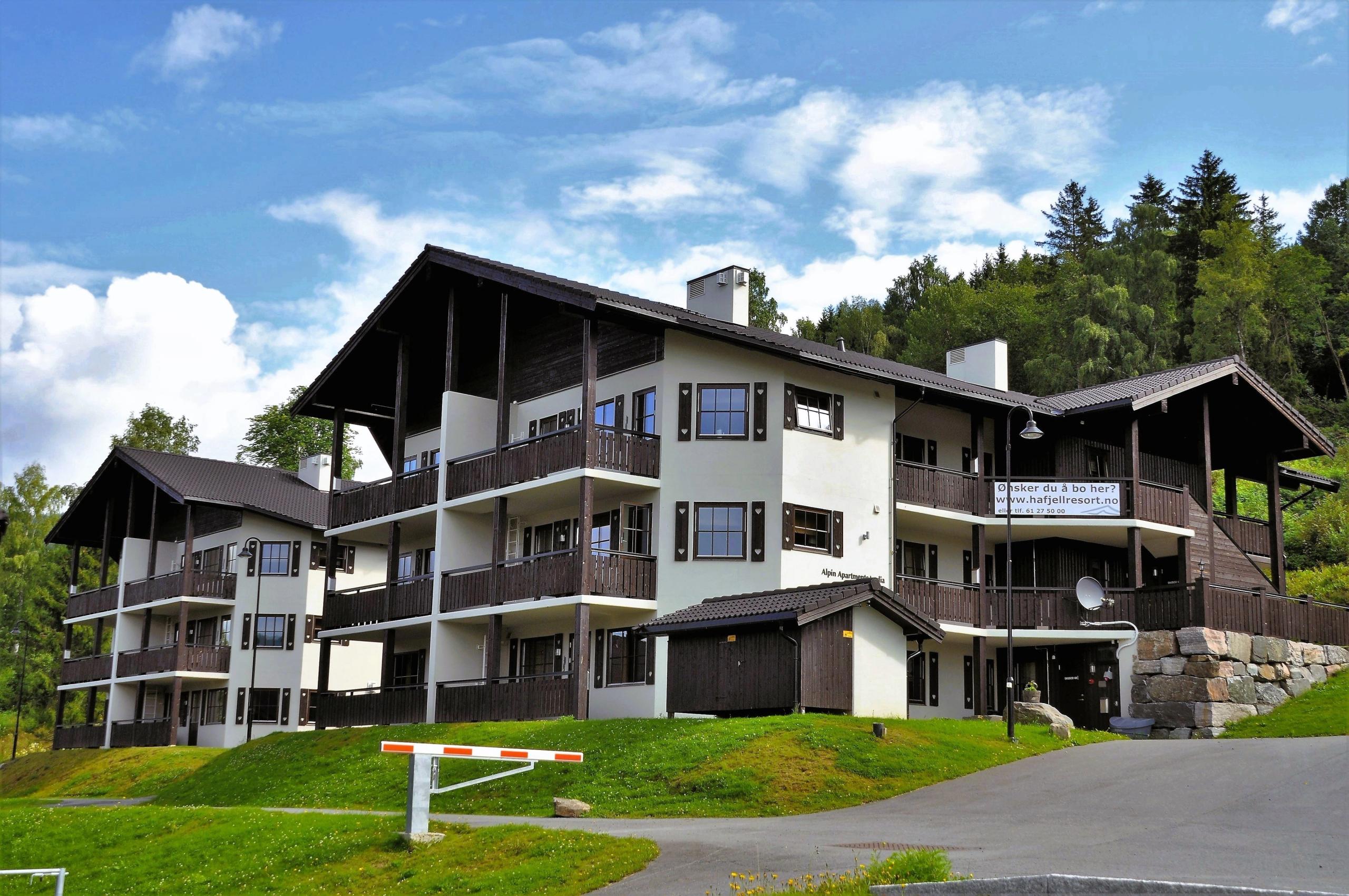 Stort leilighetsbygg med tre- og hvit fasade og balkonger, ved grønn bakke, skog og blå himmel..