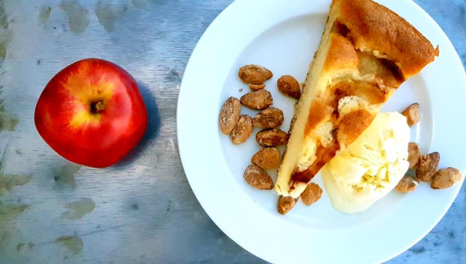 A red apple sits next to a white plate with a slice of apple cake, a scoop of vanilla ice cream, and caramelized almonds. The colors are bright and fr