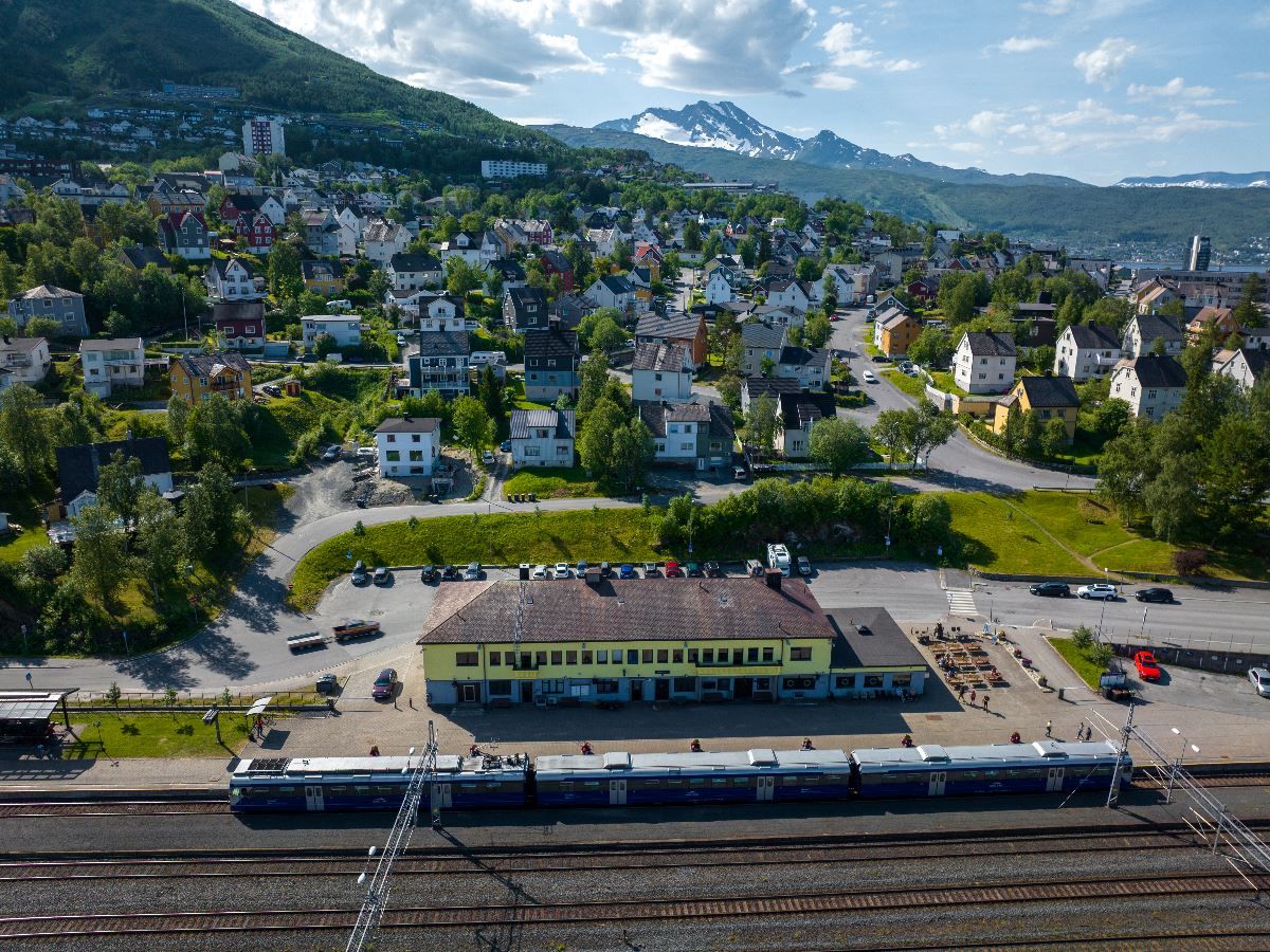 Luftfoto av togstasjon med tog og spor foran en by med hus i åsside, grønt landskap og snødekte fjell i bakgrunnen..