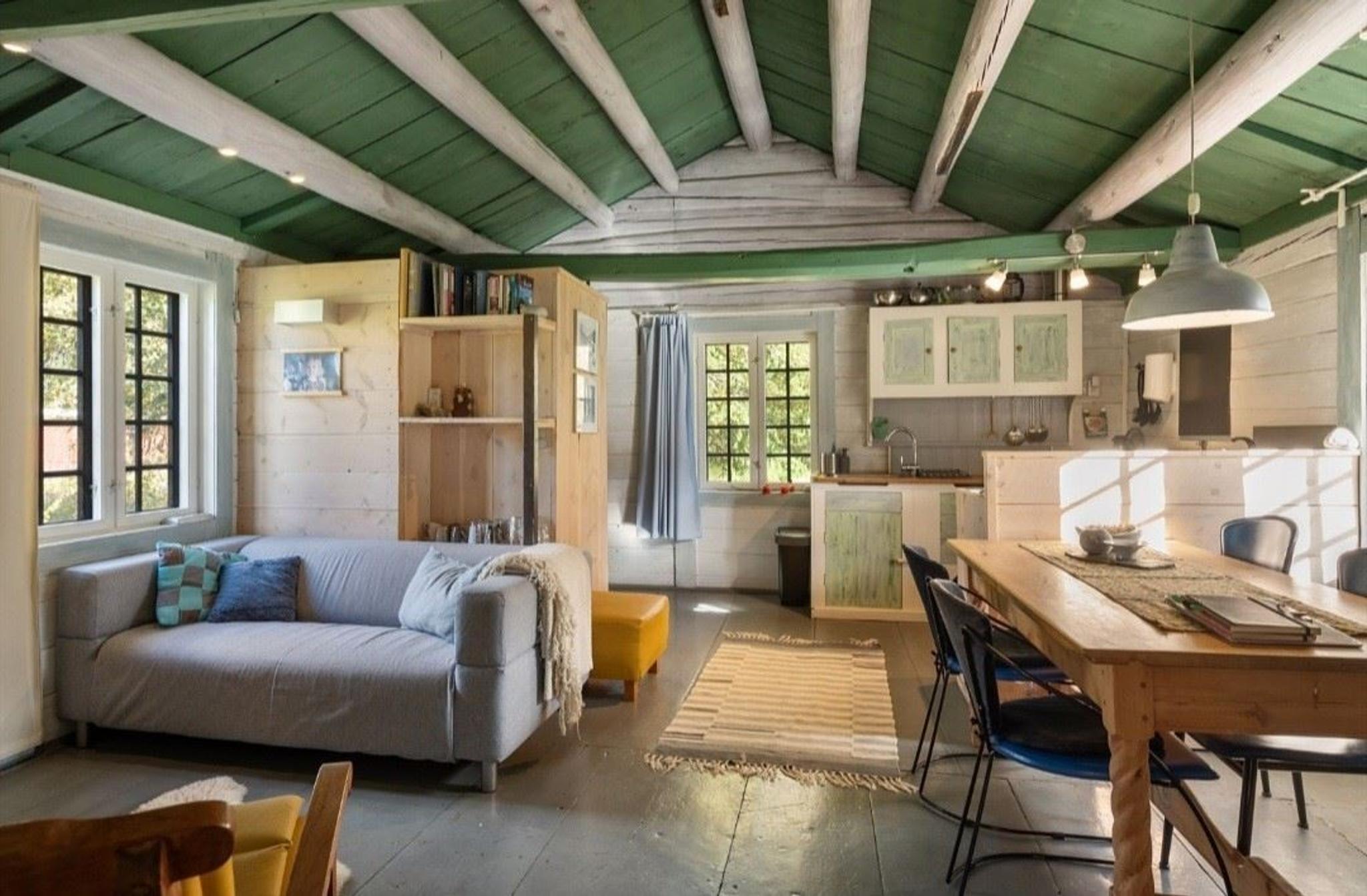 Indoor photo of a bright living room and kitchen with wooden floors, ceiling beams, and large windows letting in natural light.