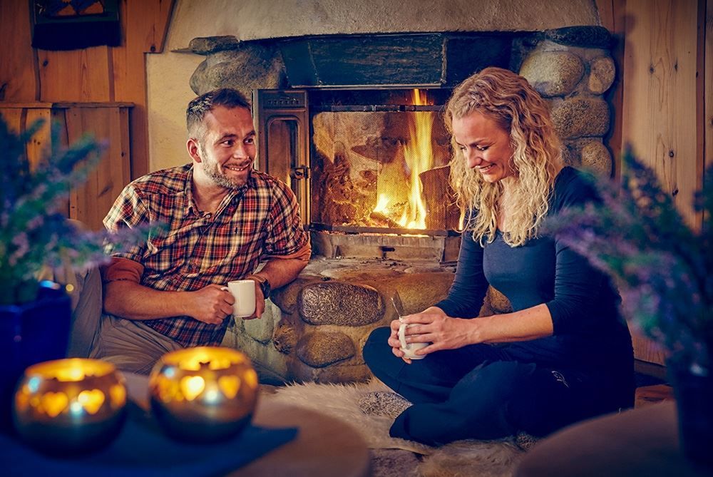 Two people sit indoors by a stone fireplace with a burning fire, holding mugs in a warm wooden cabin room..