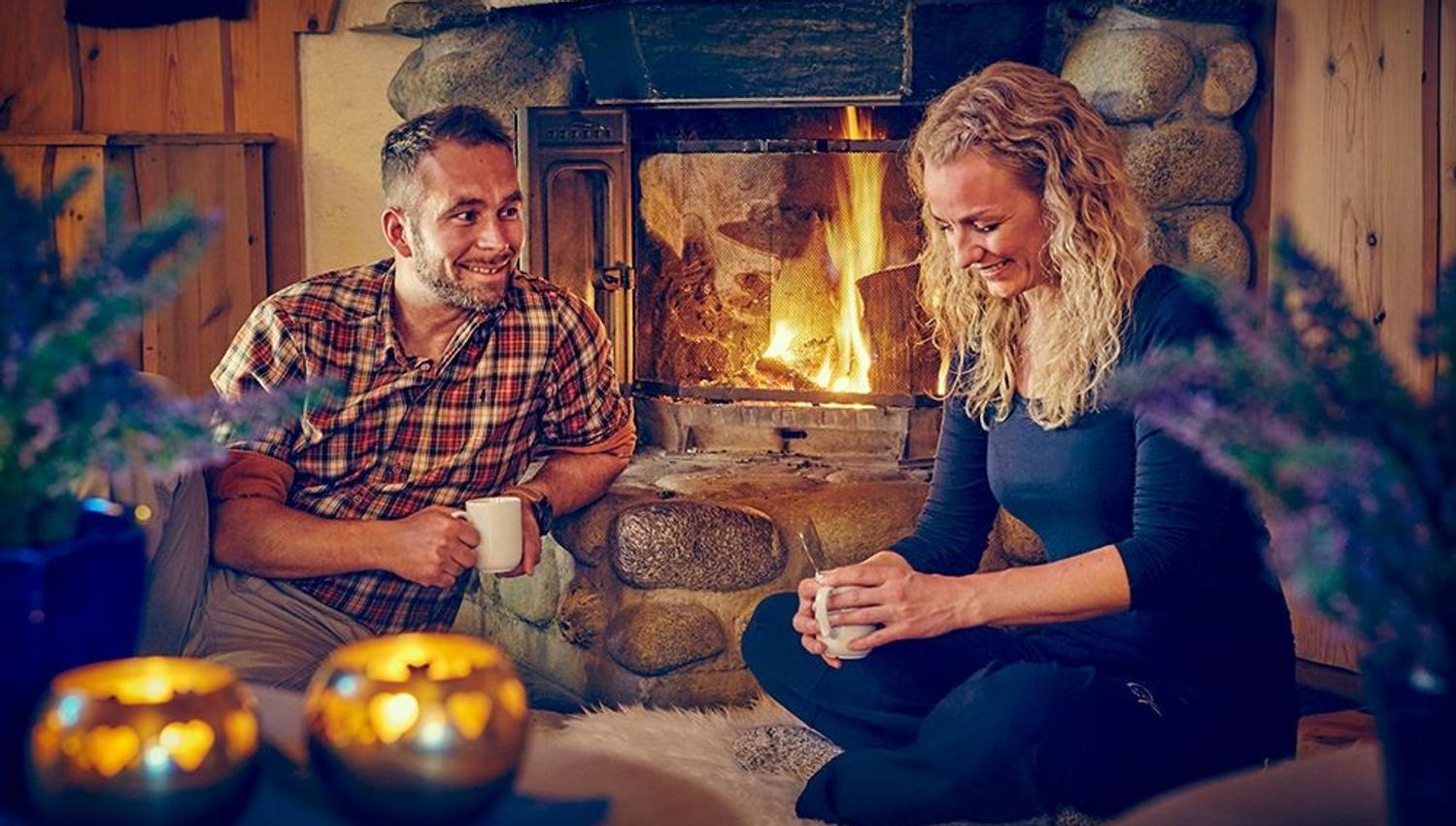 Two people sit indoors by a stone fireplace with a burning fire, holding mugs in a warm wooden cabin room..