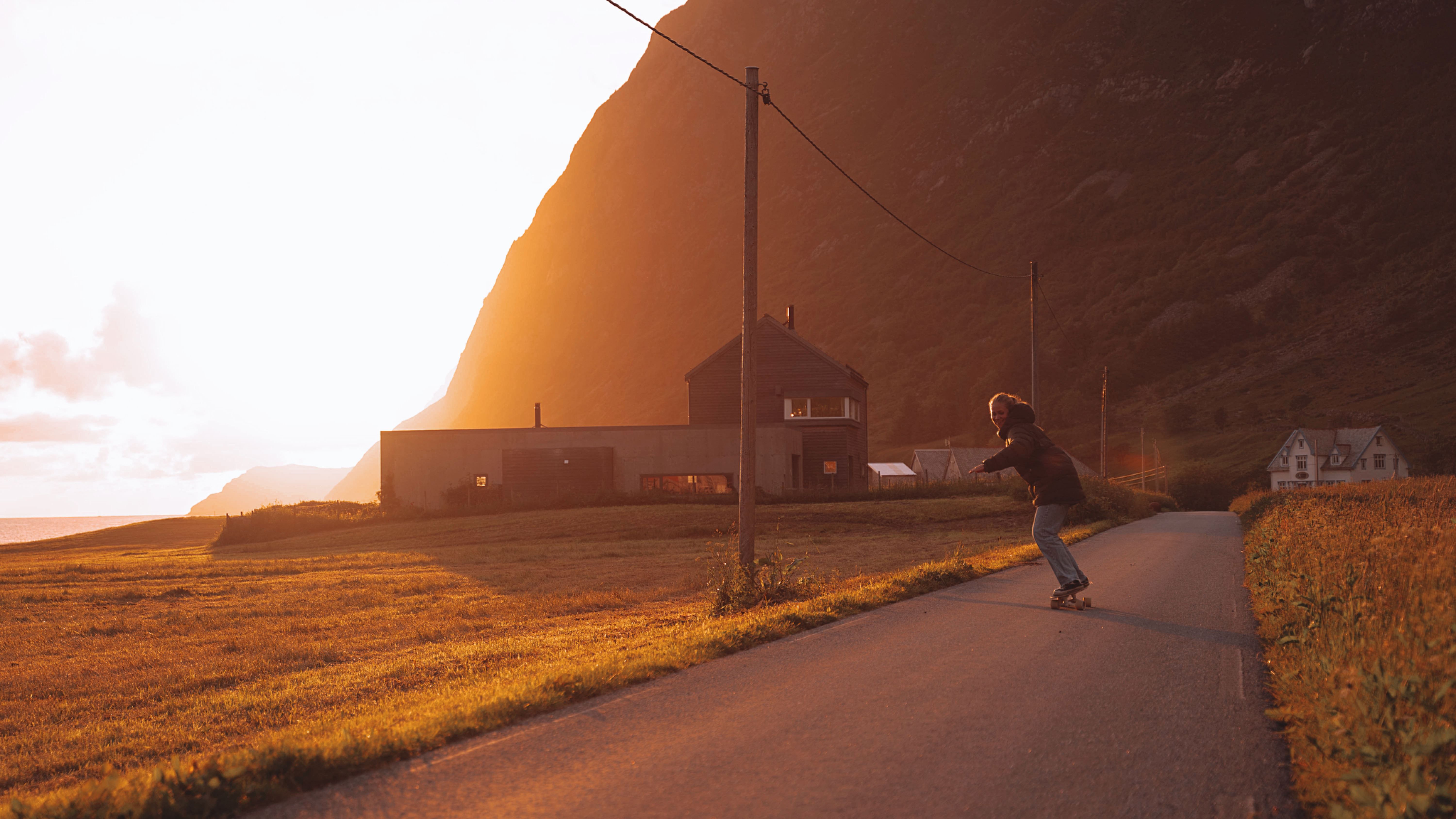 A person on a skateboard rides a narrow road past small houses and poles, with sunlit mountains in the background.
