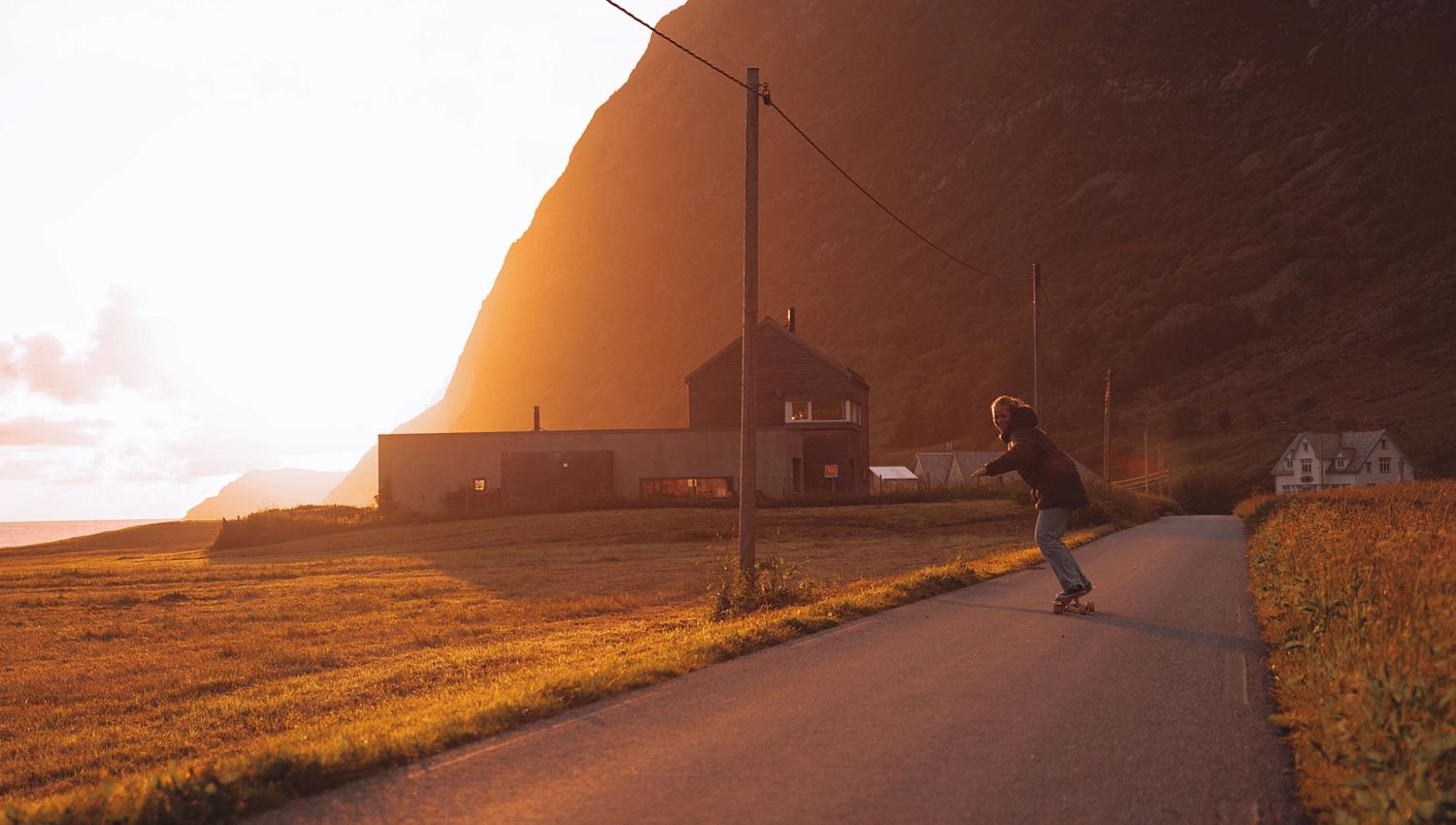 A person on a skateboard rides a narrow road past small houses and poles, with sunlit mountains in the background.