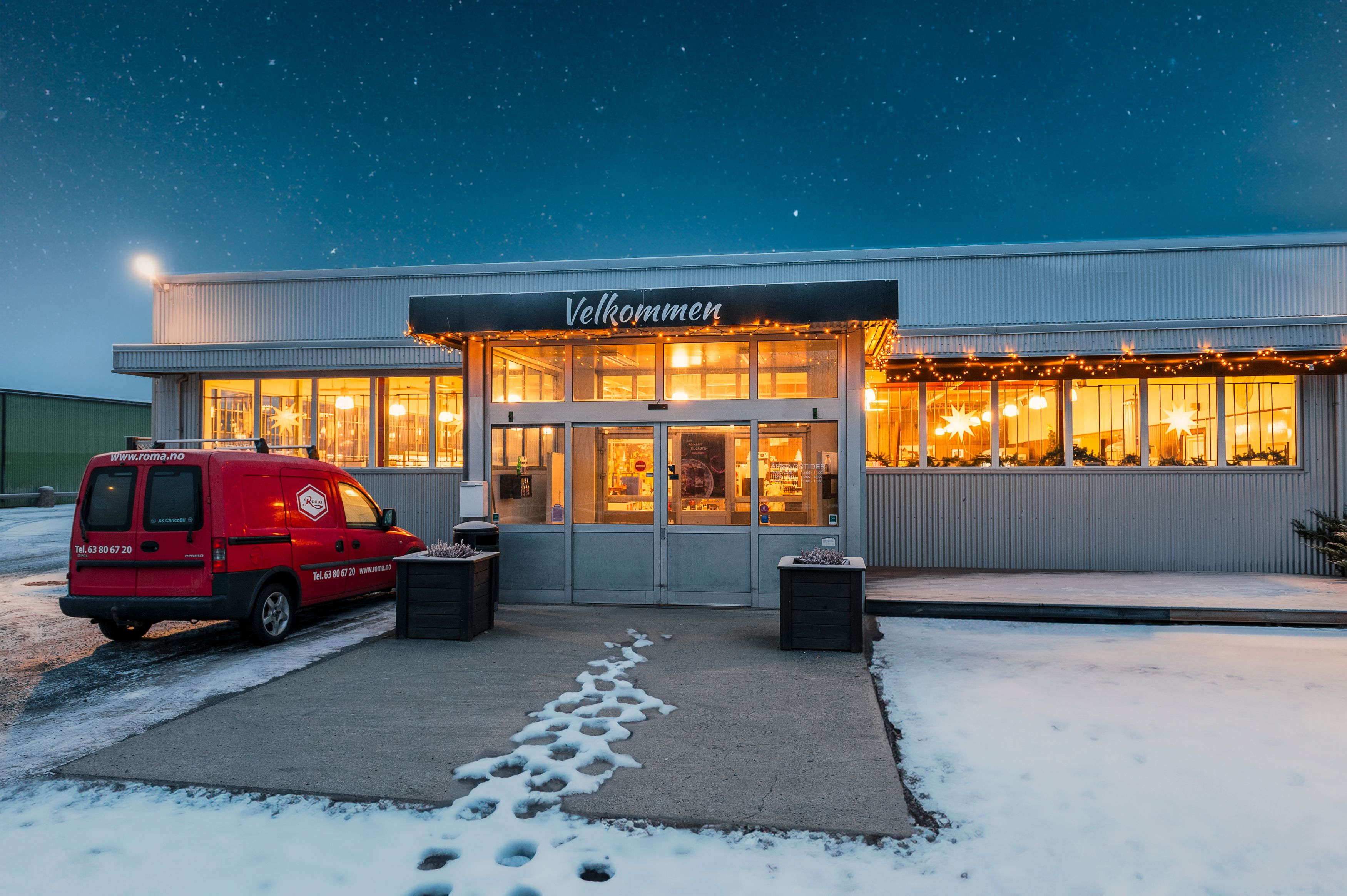 Butikkinngang med skiltet «Velkommen», julelys i vinduene og snø på bakken. Rød varebil står parkert foran.