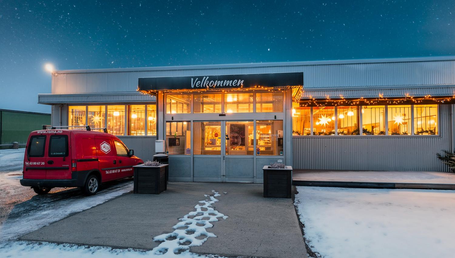 Butikkinngang med skiltet «Velkommen», julelys i vinduene og snø på bakken. Rød varebil står parkert foran.