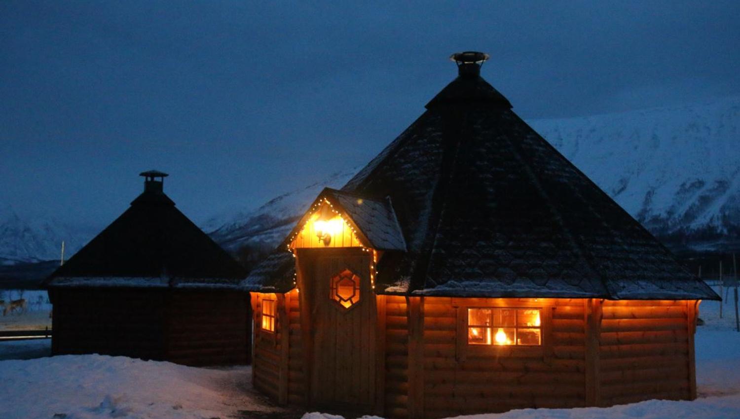 Opplyst tretakshytte i snødekt landskap i mørket.