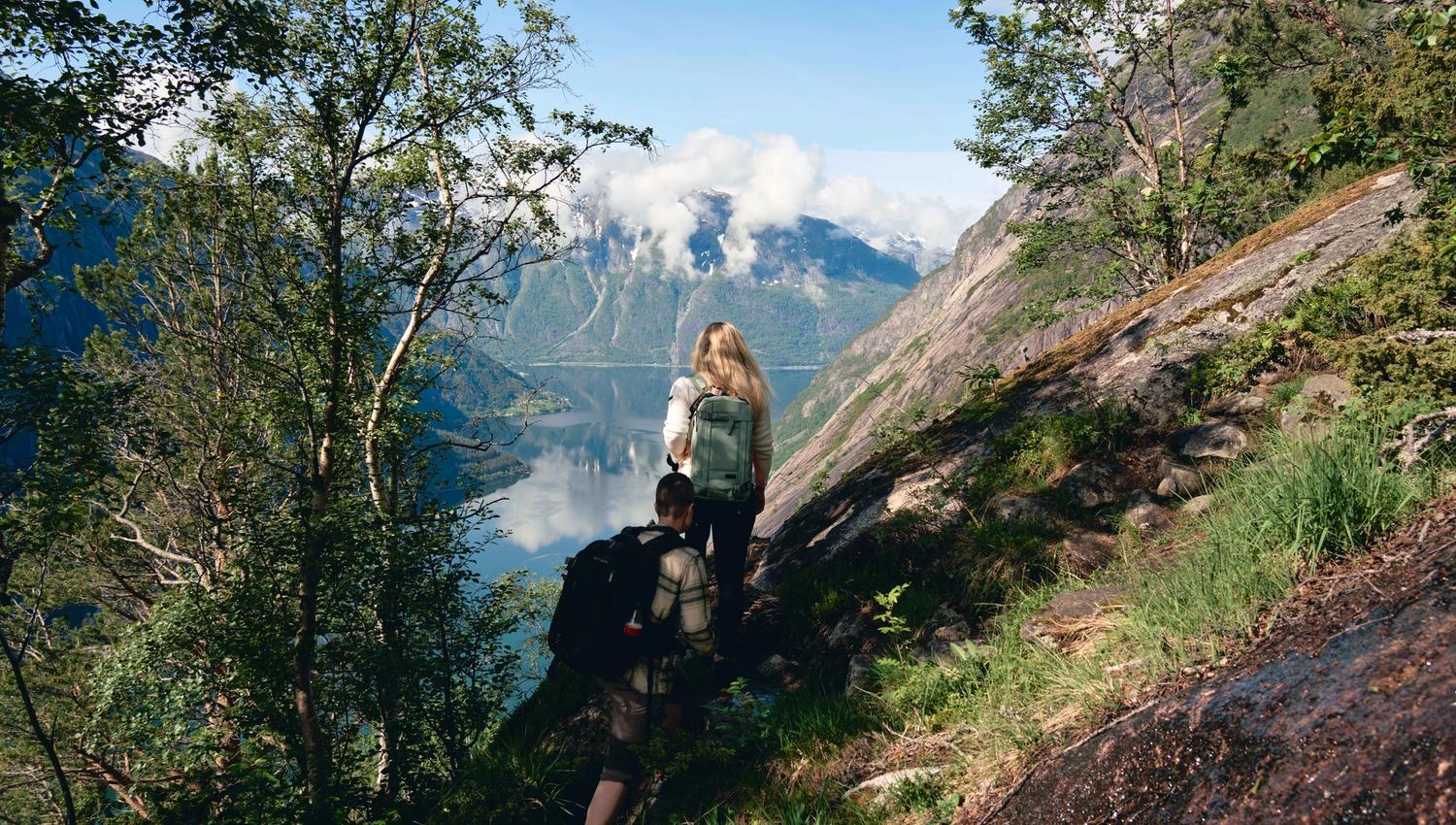 Two women hiking the steep Kjeåsen path, with the fjord and mountains in the background.