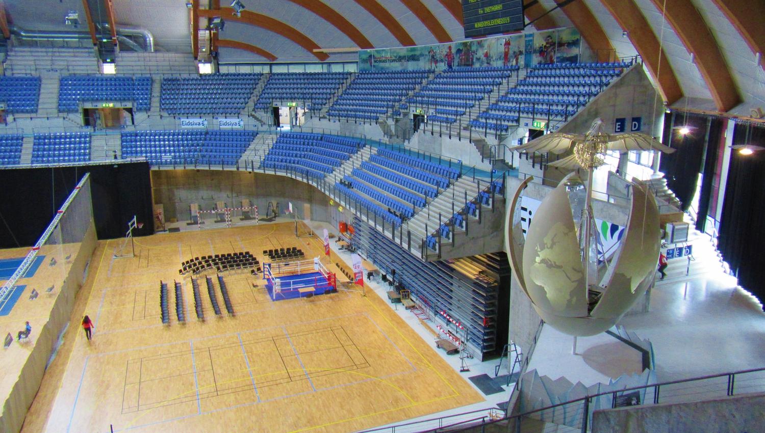 Stor innendørs idrettshall med blå tribuneseter, trebane og en stor dekorativ globeskulptur ved siden..