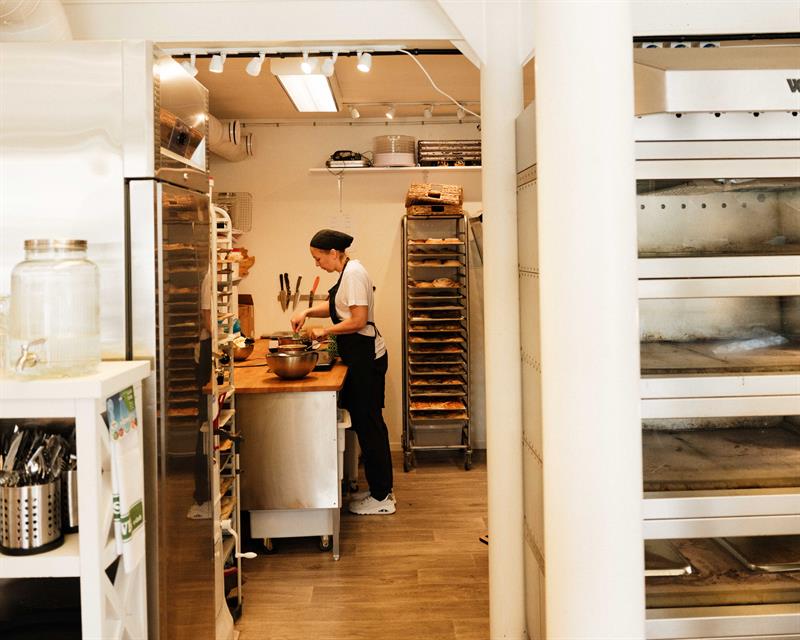A baker works attentively at the counter inside Iversen Bakery and Gallery, with dough, baking trays and racks filled with pastries around her.
