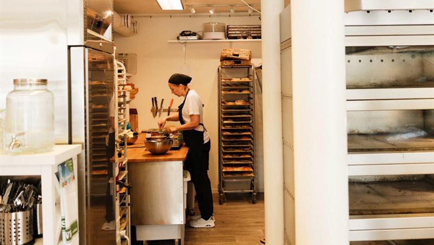A baker works attentively at the counter inside Iversen Bakery and Gallery, with dough, baking trays and racks filled with pastries around her.