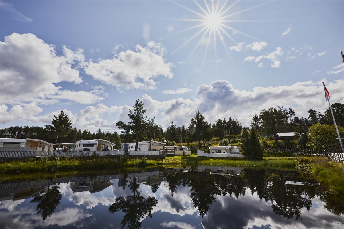 Campingplassen i strålende sol med noen få skyer på himmelen. Viser blikkstille vann i forkant av campingplassen.